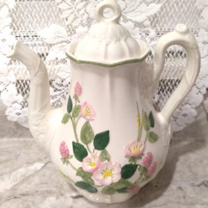 May include: Cream-colored ceramic teapot with a floral design. The teapot features a curved spout and handle, a lid with a ring, and is decorated with pink flowers, green leaves, and pink buds. The rim of the lid is outlined in green.