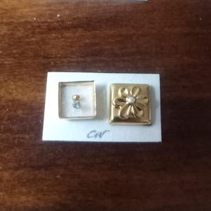 May include: Two gold-toned earrings on a white card. One is a small square with a clear stone and a gold stud. The other is a rectangular earring with a flower design and a pearl center. The initials "CW" are written on the card.