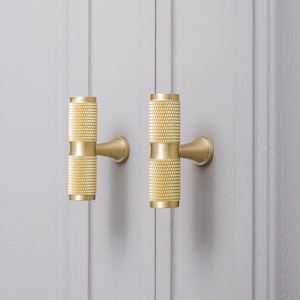 Pode incluir: Dois botões de armário dourados com um design cilíndrico texturizado. Os botões estão montados em uma parede cinza claro.