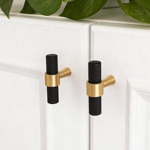 May include: Two black and gold cabinet pulls. The cylindrical black handles have a textured surface and are accented with a gold-colored ring and a gold-colored arm. The pulls are mounted on a white cabinet door.