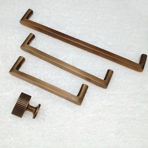 Op de afbeelding: Drie antieke messing kastgrepen met een vierkante vorm en een afgeronde rand. De grepen zijn in verschillende maten. Er is ook een kleine antieke messing knop met een geribbeld ontwerp.