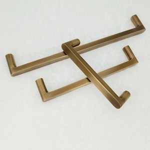 Può includere: Tre maniglie per armadio in ottone antico con forma rettangolare e bordi arrotondati. Le maniglie sono disposte a croce.
