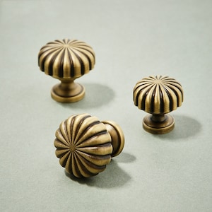 May include: Three antique brass cabinet knobs with a fluted, sunburst design. The knobs have a round, domed top and a short, cylindrical base.