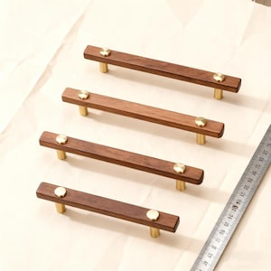 May include: Four wooden cabinet pulls with brass accents. Each pull is rectangular, with a dark brown finish and a circular brass accent on each end. The pulls are mounted on brass posts. A metal ruler is in the background.