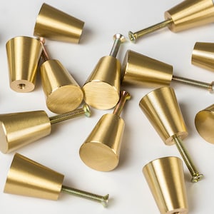 May include: A collection of brass cabinet knobs with a conical design. Each knob has a flat, circular base and a threaded screw for easy installation. The knobs are a warm gold color and are scattered on a white surface.