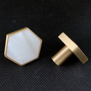 Może przedstawiać: Dwa sześciokątne gałki do szafki z mosiężnym wykończeniem. Jedna gałka ma białe centrum, a druga jest z litego mosiądzu.