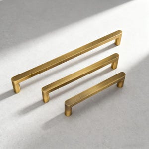 Könnte beinhalten: Drei messingfarbene Schrankgriffe in verschiedenen Längen, in einer Reihe angeordnet. Die Griffe haben eine rechteckige Stangenform mit abgerundeten Enden. Sie sind vor einem hellgrauen Hintergrund platziert. Möbelbeschläge.