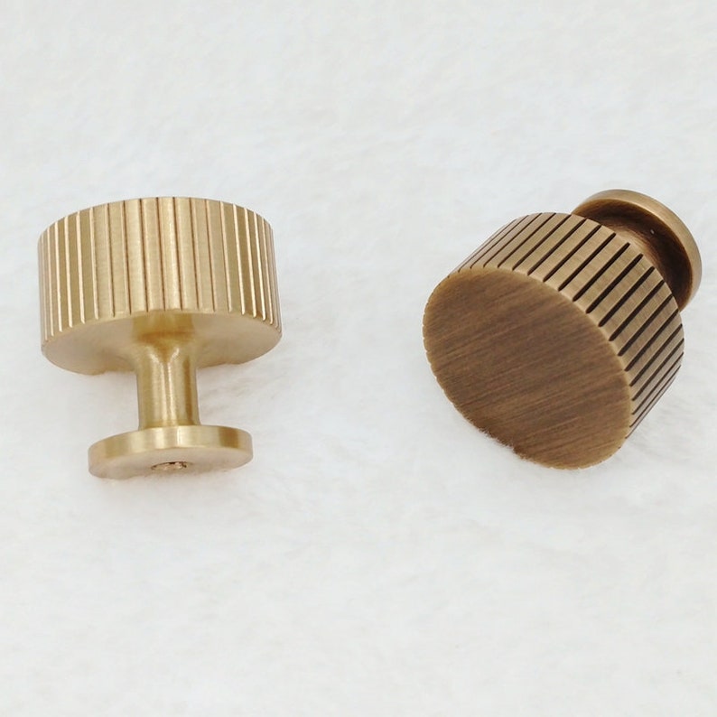 May include: Two gold-toned cabinet knobs with a ribbed design. The knobs have a round base and a cylindrical stem.