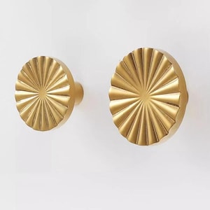 May include: Two gold-toned metal wall hooks with a sunburst design. The hooks are round and have a raised, textured surface.