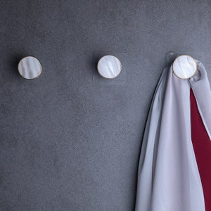 Peut inclure: Trois crochets ronds blancs avec une garniture dorée montés sur un mur gris. Une écharpe grise et rouge est accrochée à l'un des crochets.