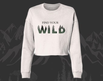 May include: White cropped sweatshirt with long sleeves featuring a forest green graphic of trees forming the word "WILD" with the text "FIND YOUR" above it.
