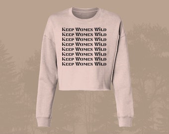 May include: A light pink cropped sweatshirt with long sleeves and the text "KEEP WOMEN WILD" repeated eight times in black.