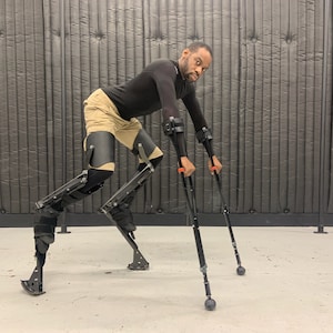 May include: A pair of black mechanical leg stilts with adjustable straps and curved foot bases, paired with two black forearm crutches featuring orange accents. The equipment is designed for mobility, featuring rigid leg supports and metal frames.