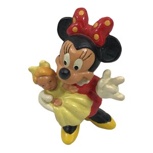 May include: A red and black figurine of Minnie Mouse holding a yellow-dressed doll. Minnie Mouse is wearing a red dress with white polka dots and a red bow with yellow polka dots.