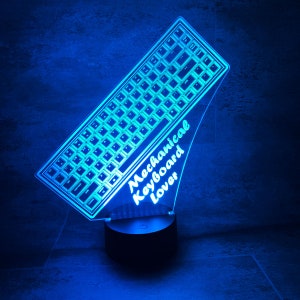Könnte beinhalten: Eine blau beleuchtete LED-Lampe in Form einer mechanischen Tastatur. Die Lampe hat einen schwarzen Sockel mit einem Ein-/Ausschalter. Die Worte "Mechanical Keyboard Lover" stehen auf der Seite der Tastatur.