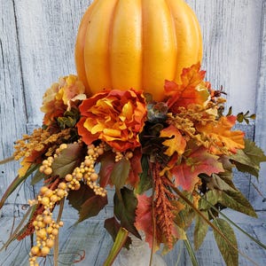 May include: A fall-themed centrepiece featuring a large orange pumpkin, surrounded by a variety of autumn foliage, including red, orange, and yellow leaves, and small yellow berries. The centrepiece is on a white pedestal.