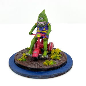 Puede incluir: Figura de un cíclope verde montado en un triciclo rojo. La criatura lleva un atuendo morado y amarillo. La base es marrón con follaje amarillo, sobre una base circular azul. La figura tiene un diseño caprichoso y de dibujos animados.