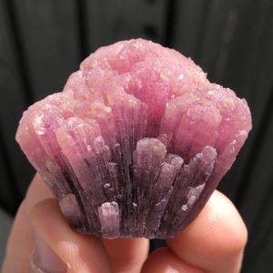 May include: A close-up of a pink tourmaline crystal cluster. The crystal has a layered appearance with a gradient of colour, transitioning from a deep magenta at the base to a lighter pink at the top. The surface has a rough, textured appearance.