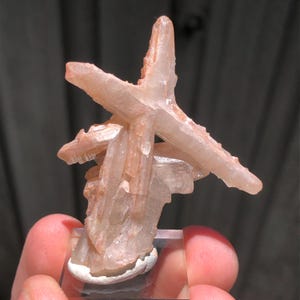Puede incluir: Se muestra un grupo de formaciones cristalinas de color melocotón, que se asemejan a una estrella. Los cristales son translúcidos con una textura ligeramente rugosa. La base es un bloque rectangular transparente con una base blanca y redondeada. El fondo es borroso y de tono neutro.