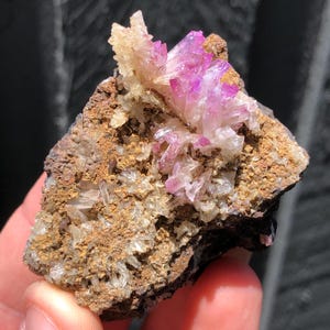 May include: A close-up of a mineral specimen featuring clusters of vibrant, crystalline formations. The crystals display a gradient of colors, from pale yellow to deep magenta, set against a rough, brown matrix. The specimen is held in a hand.