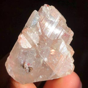 May include: A translucent, pale pink mineral specimen with a crystalline structure. The stone features sharp, angular facets and visible striations on its surface, with small, dark, metallic-looking inclusions embedded within the light-coloured crystal.