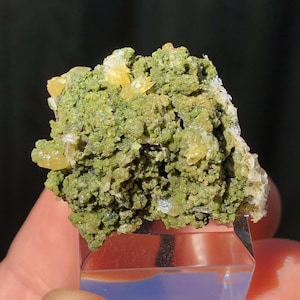 May include: A cluster of green and yellow crystals. The crystals are arranged in a random pattern and have a rough, textured surface. The crystals are on a clear display stand.
