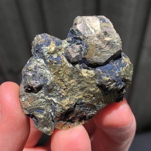 May include: A natural pyrite specimen, often called fool's gold, with a metallic sheen. The rock displays gold, bronze, and dark blue-grey hues. The pyrite is held in a hand, revealing its uneven form and texture. Measurements are not available.