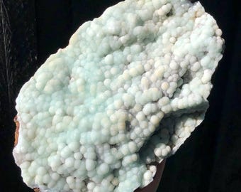 Op de afbeelding: Een onregelmatig gevormd mineraal specimen met een oppervlak bedekt met kleine, afgeronde, lichtblauwe en witte formaties. De basis heeft een donkerbruine, ruwe textuur. De algehele uitstraling is organisch en natuurlijk.