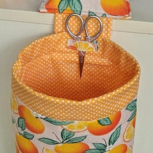 May include: A fabric organizer with an orange and white citrus fruit pattern, holding a pair of small scissors. A matching pin cushion with pins is visible. The interior of the organizer is orange with white polka dots.