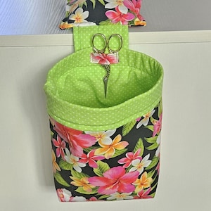May include: A fabric sewing organizer with a black floral print, a lime green interior, and a matching pin cushion. The organizer has a pocket for scissors and is designed to hang. The pin cushion has colorful pins.