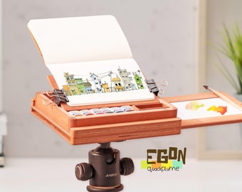 May include: A wooden artist's easel with a watercolor palette, a sketchbook, and a paintbrush. The easel is set up on a tripod and has a small tray for holding paints. The sketchbook is open to a page with a drawing of a city.