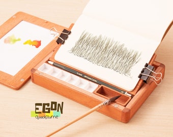 May include: A wooden artist's easel with a watercolor palette, a pencil, a paintbrush, and a sketchbook with a drawing of grass. The easel is open and the sketchbook is propped up at an angle. The easel has a small frame on the side for displaying artwork.