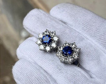 May include: A pair of flower-shaped earrings. Each earring has a large, round, deep blue gemstone at the centre, encircled by smaller, clear gemstones. The earrings are set in a silver-coloured metal, creating a sparkling, elegant design.