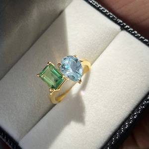 Könnte beinhalten: Ein goldener Ring mit einem birnenförmigen hellblauen Edelstein und einem quadratischen grünen Edelstein.