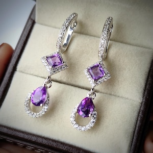 Peut inclure: Une paire de boucles d'oreilles pendantes en argent avec une améthyste violette de taille carrée entourée de petits cristaux blancs, suspendue au-dessus d'une améthyste violette en forme de poire dans un cadre circulaire de cristaux.