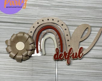 May include: A brown and white rainbow cake topper with the word "one" and the word "derful" in red. The topper is decorated with a brown flower.