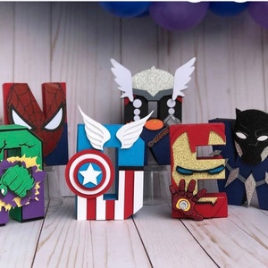 Pode incluir: Letras de cartão coloridas que soletram a palavra "NEW" com temas de super-heróis. As letras apresentam designs inspirados em Homem-Aranha, Thor, Capitão América, Homem de Ferro, Hulk e Pantera Negra. Balões visíveis no fundo.
