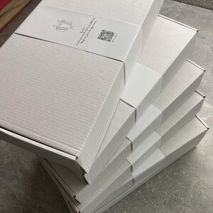 Peut inclure: Une pile de bo&icirc;tes en carton blanches, chacune avec une bande blanche autour. Les bo&icirc;tes sont rectangulaires et ont une surface textur&eacute;e. Une &eacute;tiquette avec du texte et un code QR est visible sur la bo&icirc;te sup&eacute;rieure.