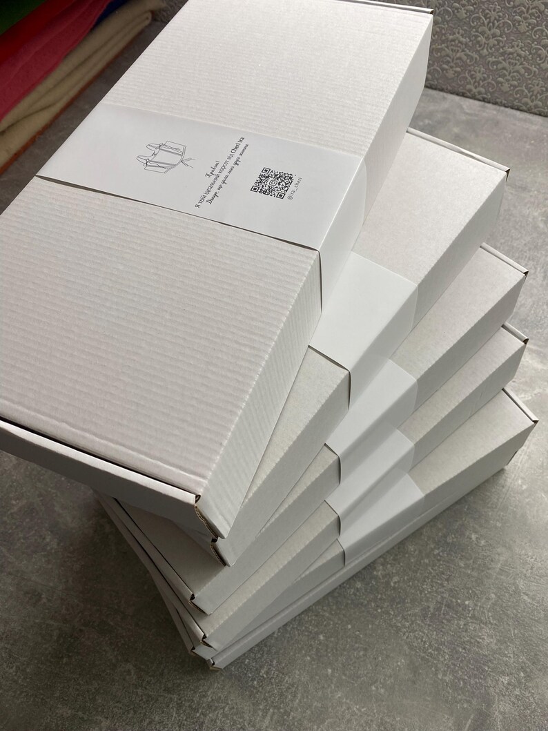 Peut inclure: Une pile de bo&icirc;tes en carton blanches, chacune avec une bande blanche autour. Les bo&icirc;tes sont rectangulaires et ont une surface textur&eacute;e. Une &eacute;tiquette avec du texte et un code QR est visible sur la bo&icirc;te sup&eacute;rieure.