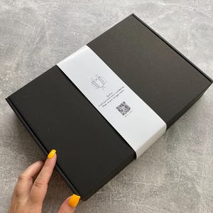 Peut inclure: Une bo&icirc;te rectangulaire noire avec une bande blanche. La bande pr&eacute;sente une illustration minimaliste et du texte. Une main avec du vernis &agrave; ongles jaune est visible.