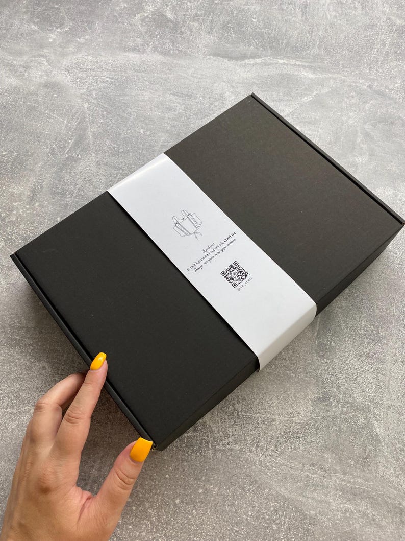 Peut inclure: Une bo&icirc;te rectangulaire noire avec une bande blanche. La bande pr&eacute;sente une illustration minimaliste et du texte. Une main avec du vernis &agrave; ongles jaune est visible.