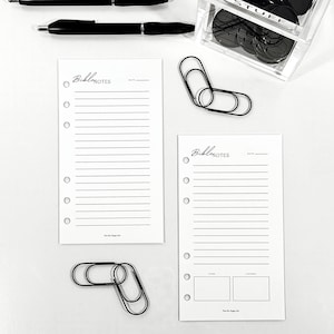 May include: An overhead shot features stationery items on a white surface. Two black pens, two black paper clips, and a clear acrylic container filled with black circular objects are visible. Two white lined paper inserts with the text "Bible Notes" are also present.