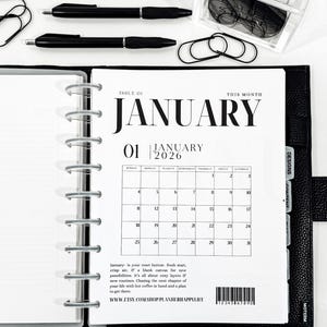May include: A black leather planner open to a January 2026 calendar page. The page includes the text "January" and a calendar grid. Black pens, paper clips, and a container of black discs are also visible.