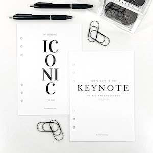 May include: A flat lay featuring stationery items. Two black pens, two white paper sheets with text, black paper clips, and a clear container filled with black circular objects. The text on the paper reads "ICONIC" and "SIMPLICITY IS THE KEYNOTE".