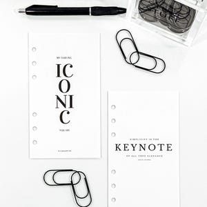 May include: A collection of office supplies including black pens, paper clips, and a clear container of black circular objects. Two white planner inserts with text that reads "ICONIC YOU ARE" and "SIMPLICITY IS THE KEYNOTE".