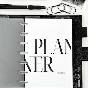 Könnte beinhalten: Schwarzer Lederplaner mit dem Wort „PLANNER“ in großen, fetten Buchstaben. Der Planer enthält transparente Trennwände, schwarze Stifte, Büroklammern und eine Brille. Der Planer ist auf einer Seite mit den Worten „MONTHLY & WEEKLY“ geöffnet.
