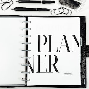 May include: Open black planner with the word "PLANNER" in large, bold, black letters. The planner is open to a white page with the text "MONTHLY & WEEKLY DESIGNED BY SARA PLAN HER HAPPY LIFE". Two black pens, paper clips, and a small container of glasses are also visible.