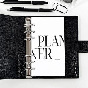 Puede incluir: Un planificador de cuero negro con anillos plateados, una inserción blanca que dice "PLANNER" y dos bolígrafos negros. También se ve un recipiente transparente con accesorios negros y un clip negro.