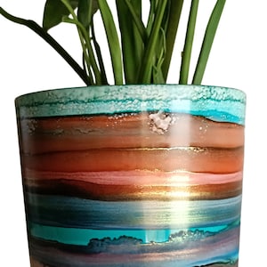 May include: A cylindrical ceramic plant pot featuring horizontal bands of colour including turquoise, copper, brown, and deep purple. The pot sits on a white saucer and contains a green leafy plant. The surface has a glossy, fluid ink finish.