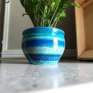 May include: A round ceramic plant pot featuring a horizontal gradient of blue, turquoise, and white colours with a glossy finish. The surface displays layered bands of varying shades of blue, creating a fluid, wave-like pattern around the exterior.